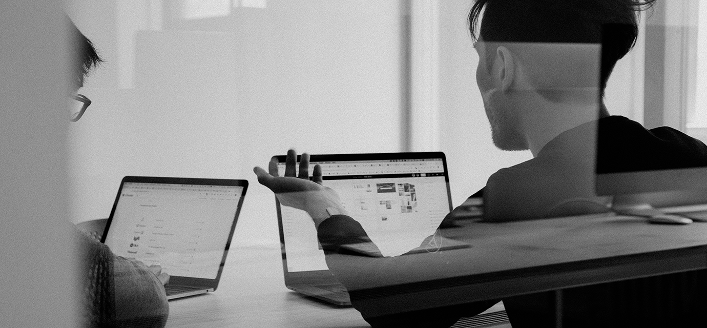 A black and white photograph of two men sitting at a desk in front of laptops.